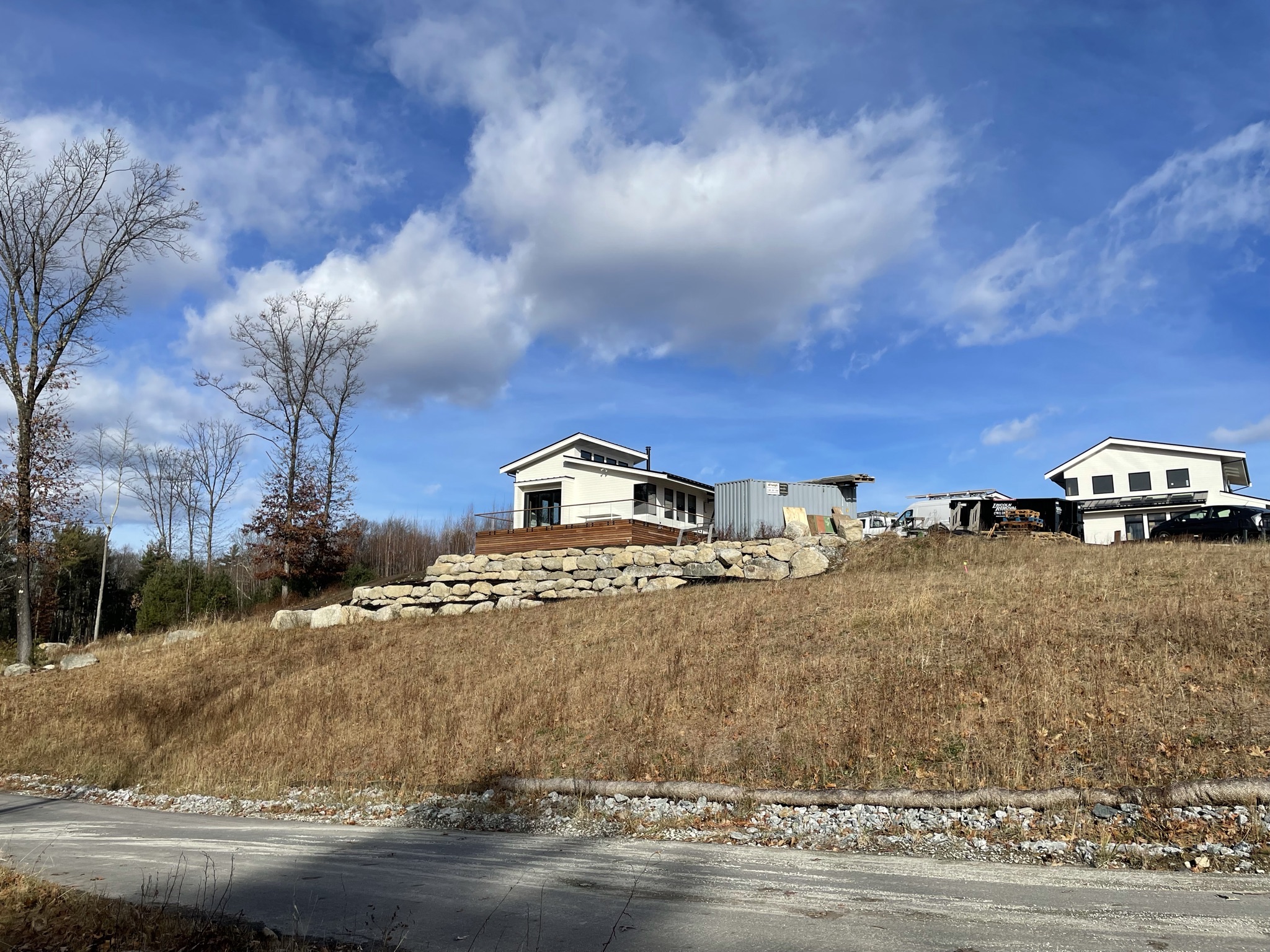 Modern home under construction sit atop a sloped, grassy hillside, with a large stacked-stone retaining wall and wooden deck in the foreground and a coir erosion-control log installed along the base of the slope beside a gravel road.